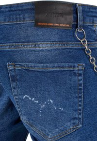 Zila denim bikses ar aizmugures kabatu, oranžām šuvēm, melnu birku ar uzrakstu "2Y jeans" un sudraba ķēdes aksesuāru.