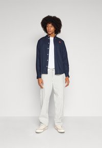 Navy button-up shirt with a chest pocket and red logo, white t-shirt underneath, paired with white striped pants and cream sneakers.