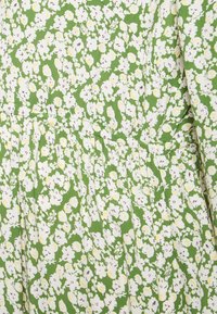 Vestido floral com um fundo verde e flores brancas e amarelas, feito de um tecido suave, com detalhes franzidos na cintura e pregas.