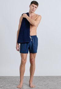 Sötétkék fürdőnadrág elasztikus derékpánttal és húzózsinórral, sötét törölközővel az egyik vállon, férfi modell által viselve.