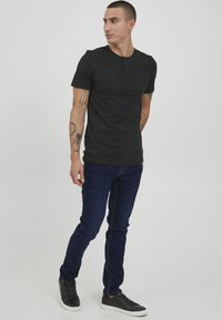 Crna kratka majica s kratkim rukavima i tri gumba, u kombinaciji s tamnoplavim trapericama i crnim tenisicama, stoji naslonjena na jednostavnu pozadinu.