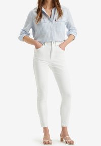 Camisa de botón a rayas azul claro con dos bolsillos en el pecho, combinada con jeans skinny blancos de tiro alto y sandalias de tacón bloque de color claro.