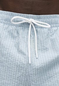 Λαστιχένια μέση με λευκό κορδόνι δεμένο σε φιόγκο σε σορτς με ανοιχτό μπλε και λευκές ρίγες σε σκουρόχρωμο δέρμα.