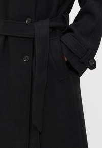 Melnas villānas mētelis ar jostu viduklī, pogu aizdari, sānu kabatām un strukturētām piedurknēm ar pogu akcentiem.