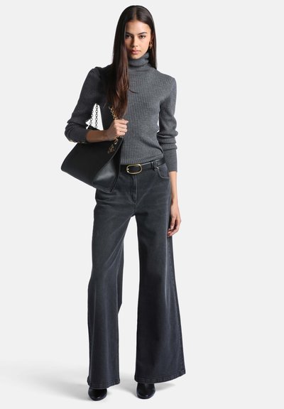 Femme portant un pull à col roulé côtelé gris et un jean noir à jambes larges, tenant un grand sac à main noir avec une bandoulière chaîne dorée.