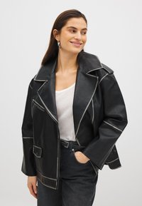 Veste en simili cuir noir avec des coutures blanches contrastantes, un col oversized, une poche zippée et des poches à rabat à l'avant.