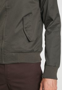 Giubbotto bomber verde scuro con chiusura a zip, polsini e orlo a coste, e una tasca laterale con patta dotata di dettaglio a bottone. Texture liscia.