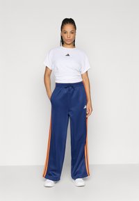 Hvit avkortet t-skjorte med svart logo, kombinert med blå treningsbukser med oransje sidestriper og snøring i midjen. Hvite sko.