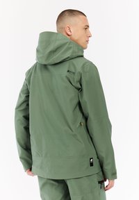 Man in een groene waterdichte jas met capuchon en bijpassende broek, gezien van achter en van de zijkant op een witte achtergrond.