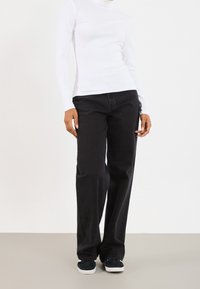 Valkoinen pitkähihainen ribbineuloksinen poolopaita yhdistettynä korkeavyötäröisiin mustiin suoralahkeisiin farkkuihin ja tummiin lenkkareihin neutraalilla taustalla.