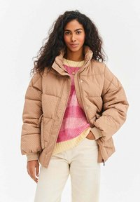 Prešívaná hnedá prešívaná bunda s vysokým golierom, zapínaním na cvoky a elastickými manžetami, nosená cez ružový a žltý sveter, s krémovými nohavicami.