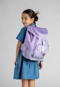 Zaino viola a pois, con striscia riflettente, spallacci regolabili e un logo. Ragazza che indossa un vestito di jeans, leggermente girata.