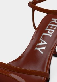 Κοντινό πλάνο καφέ δερμάτινου σανδάλου με λεπτά λουριά και λογότυπο "REPLAY" στην εσωτερική σόλα πάνω σε λευκό φόντο.