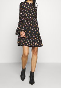 Svart blommig klänning med långa, utsvängda ärmar, som har en lager-på-lager nederkant och ett mönster av små färgglada blommor mot en mörk bakgrund.