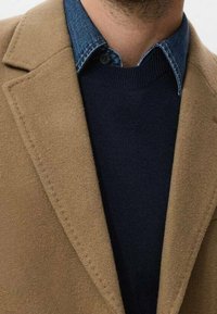 Manteau en mélange de laine couleur camel avec revers crantés, porté sur un pull bleu marine foncé et une chemise en denim. Détail de couture visible sur le bord du manteau.