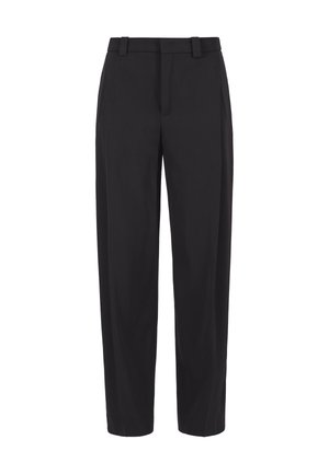 Femme portant un pantalon noir taille haute à jambes larges, un haut blanc et des chaussures noires pointues avec des nœuds, debout devant un fond uni.