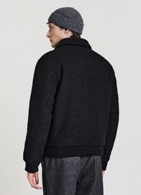 Schwarze Fleecejacke mit strukturierter Oberfläche, kurzem Kragen und gerippten Bündchen. Kombiniert mit einer grauen Strickmütze, Rückansicht.
