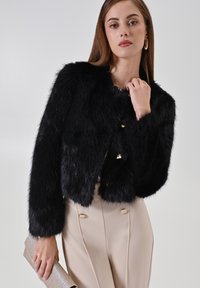 Veste courte en fausse fourrure noire avec col rond, boutons dorés et manches longues. Associée à un pantalon beige sur mesure et une pochette pailletée.