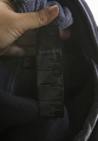 Peça de vestuário cinza com capuz e uma textura suave, apresentando uma etiqueta de cuidados em várias línguas, detalhando a composição do tecido e as instruções de lavagem.