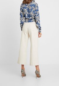 Kvinna som står i vita vida byxor, blommig blå långärmad skjorta och högklackade skinnskodon, sedd bakifrån.