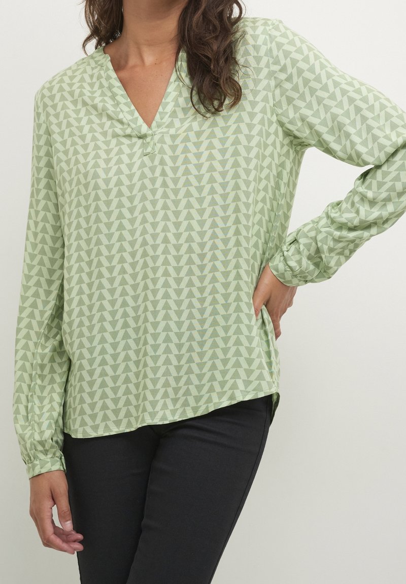 Mujer que lleva una blusa de manga larga verde claro con un patrón de triángulos geométricos, combinada con pantalones negros, de pie con una mano en la cadera.