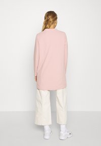 Túnica de manga larga color rosa claro, hecha de tela suave, con un cuello sencillo y un corte recto, combinada con pantalones anchos blancos.