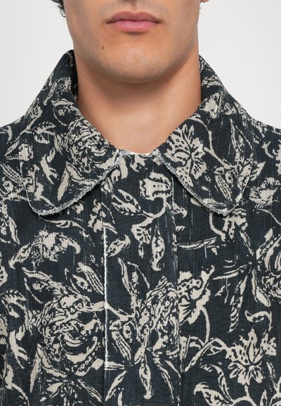 Material texturat de culoare neagră cu un model floral alb, având un guler ascuțit și un fermoar cu butoane.