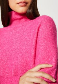 Fuchsiový pletený rolák vyrobený z měkké texturované látky, s dlouhými rukávy a uvolněným střihem. Detailní záběr na výstřih a rukáv.