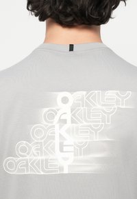 Γκρίζα βαμβακερή μπλούζα με κοντό μανίκι, η οποία διαθέτει ένα μεγάλο, στυλιζαρισμένο άσπρο λογότυπο "Oakley" στο πίσω μέρος, με κυματιστά σχέδια και μια μικρή μαύρη ετικέτα στον λαιμό.