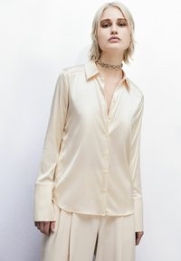Blusa in satin crema con colletto, bottoni frontali e maniche lunghe con ampi polsini. Texture liscia con finitura lucida.