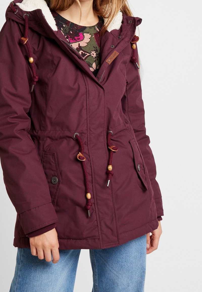 Eine Frau trägt eine lila winterliche Jacke mit Kapuze und Zugbändern sowie ein Blumenhemd darunter, kombiniert mit blauen Jeans vor einem weißen Hintergrund.