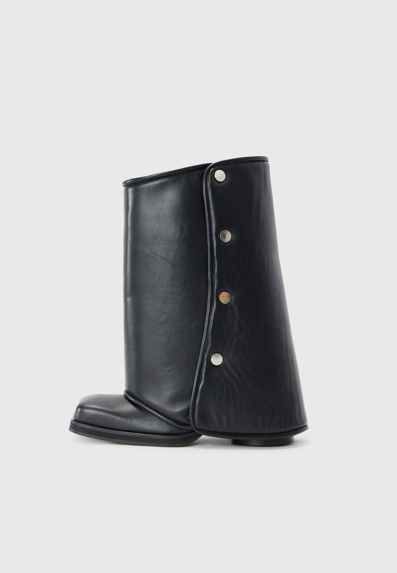 Bottine en cuir noir avec un design incurvé, dotée de fermetures à pression en argent sur le côté et d'une semelle plate et robuste. Texture lisse.