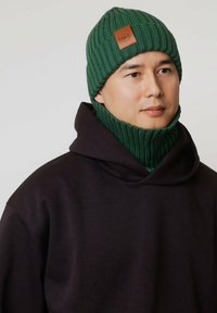 Zielona dzianinowa czapka z naszywką z logo na skórze, zestawiona z dopasowanym prążkowanym kominem. Nosi się ją na ciemnobrązowej bluzie z kapturem z szerokim kołnierzem.