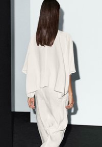 Une femme aux longs cheveux bruns porte une tenue ample de couleur gris clair, s'éloignant d'un fond minimaliste avec un éclairage doux.