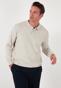 Cămașă polo lungă cu mâneci bej deschis, cu o textură ribbed, guler butonat și mansete. Purtați cu pantaloni închiși la culoare, cu mâinile în buzunare.