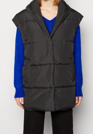 Veste noire matelassée sans manches portée sur un pull à manches longues bleu vif et un pantalon noir.