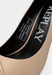 Chaussure à talon haut beige en matériau synthétique à texture lisse, avec un bout arrondi et une étiquette intérieure visible portant le nom de la marque "REPLAY".