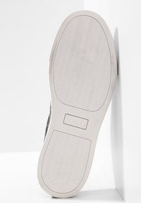 Semelle d'une chaussure blanche avec un motif ondulé et "POLO" en relief au centre, appuyée contre un mur d'angle blanc.