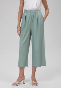 Pantalon court vert clair avec une taille élastique et des plis. Confectionné dans un tissu lisse, il comporte des poches latérales et une coupe décontractée.