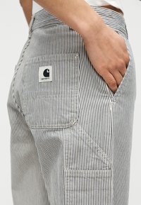 Άτομο που φοράει ριγέ παντελόνι Carhartt με το χέρι στην εμπρόσθια τσέπη, εστιάζοντας στις πίσω και πλευρικές τσέπες σε κοντινή προβολή.