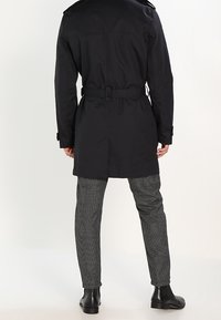 Svart trenchcoat med bälte i midjan och axelkappar, i kombination med grå hundtand mönstrade byxor och svarta ankelstövlar.