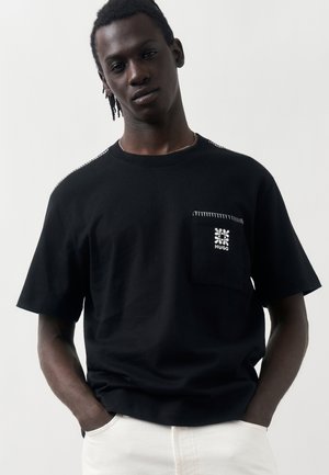 Νέος άνδρας με κοντά dreadlocks που φοράει μαύρο μπλουζάκι με λευκά ραφές και λογότυπο στην μπροστινή τσέπη, με τα χέρια στις τσέπες λευκών παντελονιών.