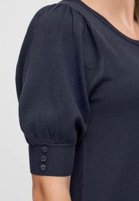 Top tricotat de culoare bleu marin, cu mâneci umflate și trei nasturi la manșetă. Textură netedă cu detalii cu anumite dungi la decolteu și la mânecă.