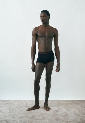 Young man standing barefoot on carpet, wearing black boxer briefs with red "HUGO" logo waistband, against plain light background.