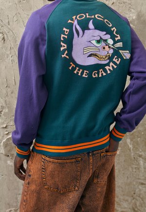 Grønne og lilla sweatshirt med et cartoon kattegrafik på ryggen, med teksten "VOLCOM PLAY THE GAME" i orange. Ribbeformede orange detaljer.