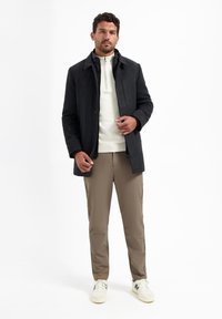 Donkergrijze jas met een kraag over een witte rits-sweater, gecombineerd met beige broek en witte sneakers. Glad stof met een getailleerde pasvorm.