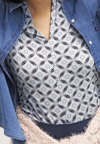 Krageblå denimjacka över en mönstrad grå skjorta med svarta geometriska former. Texturerad beige tröja med mörk midja under.