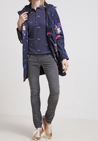 Marinblå blommig regnjacka med huva, kombinerad med en mörk polo skjorta, grå skinny jeans och beige sneakers med vita snörningar.