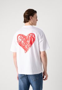 Balta kokvilnas T-kreklu ar brīvu piegriezumu, aizmugurē ar lielu sarkanu scriblētu sirds grafiku. Priekšpuse vienkārša, ar īsām piedurknēm.