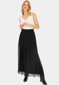 Czarna, plisowana spódnica maxi z lekkiego materiału, zestawiona z białym, bezrękawnikowym topem. Modelka stoi w czarnych, na obcasie botkach, na neutralnym tle.